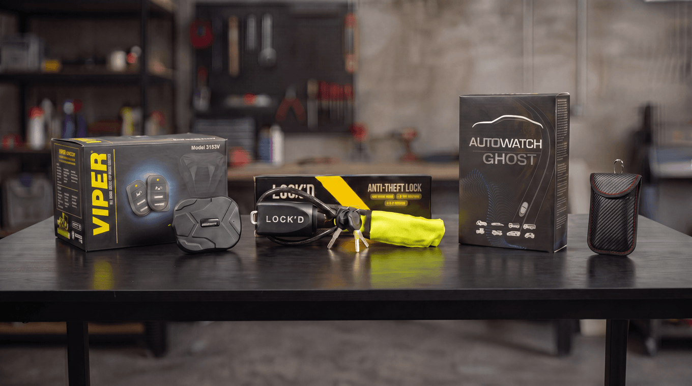 A GPS tracker, Faraday pouch, car alarm siren, Ghost immobiliser unit and a yellow Lock'd steering lock lined up on a workshop bench.