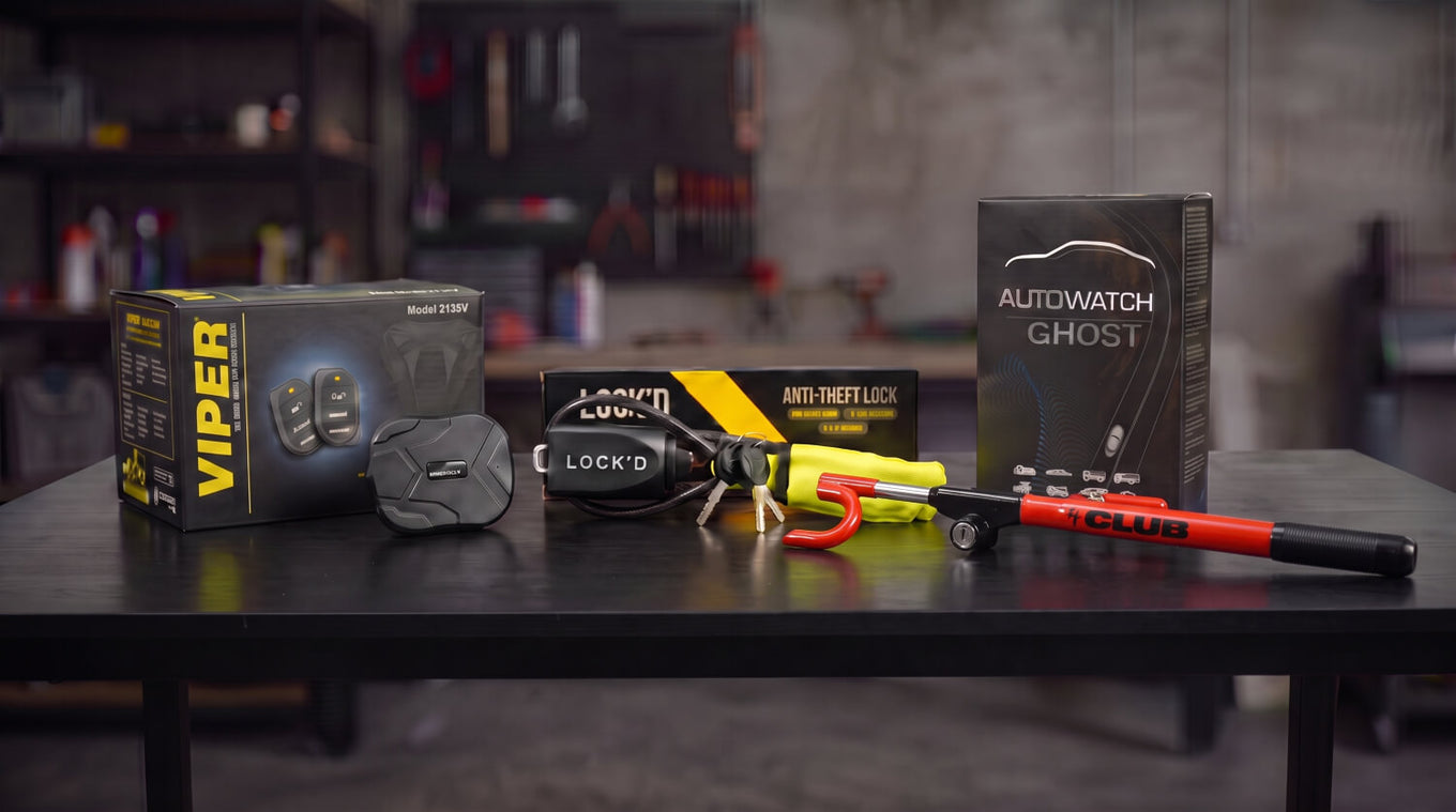 A GPS tracker, Faraday pouch, car alarm siren, Ghost immobiliser unit and a yellow Lock'd steering lock lined up on a workshop bench.